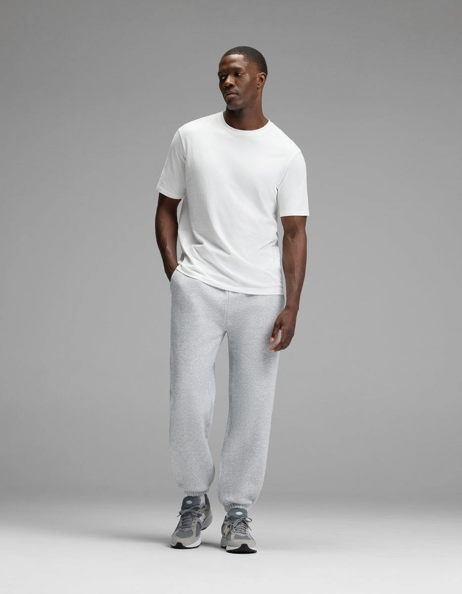 A man stands against a plain gray background, wearing a white t-shirt, Cozy Earth Men’s Cityscape 2.0 Sweatpant in light gray, and gray sneakers. His hands are in his pockets, and he is looking slightly to the side with a neutral expression.
