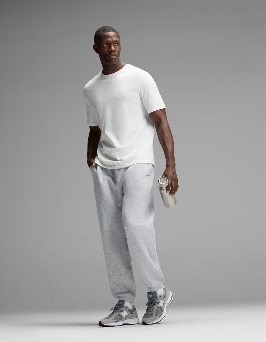 A man stands against a plain gray background, looking to the side, dressed in a white T-shirt and gray sneakers while holding a clear water bottle. He is wearing Cozy Earth Men’s Cityscape 2.0 Sweatpant.