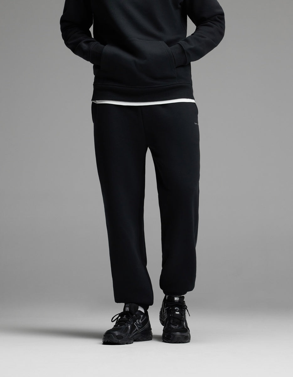 A person standing with hands in the pockets of Cozy Earth Men’s Cityscape 2.0 Sweatpant in Jet Black, paired with a black sweatshirt, white shirt underneath, and black sneakers. The upper face is not visible.