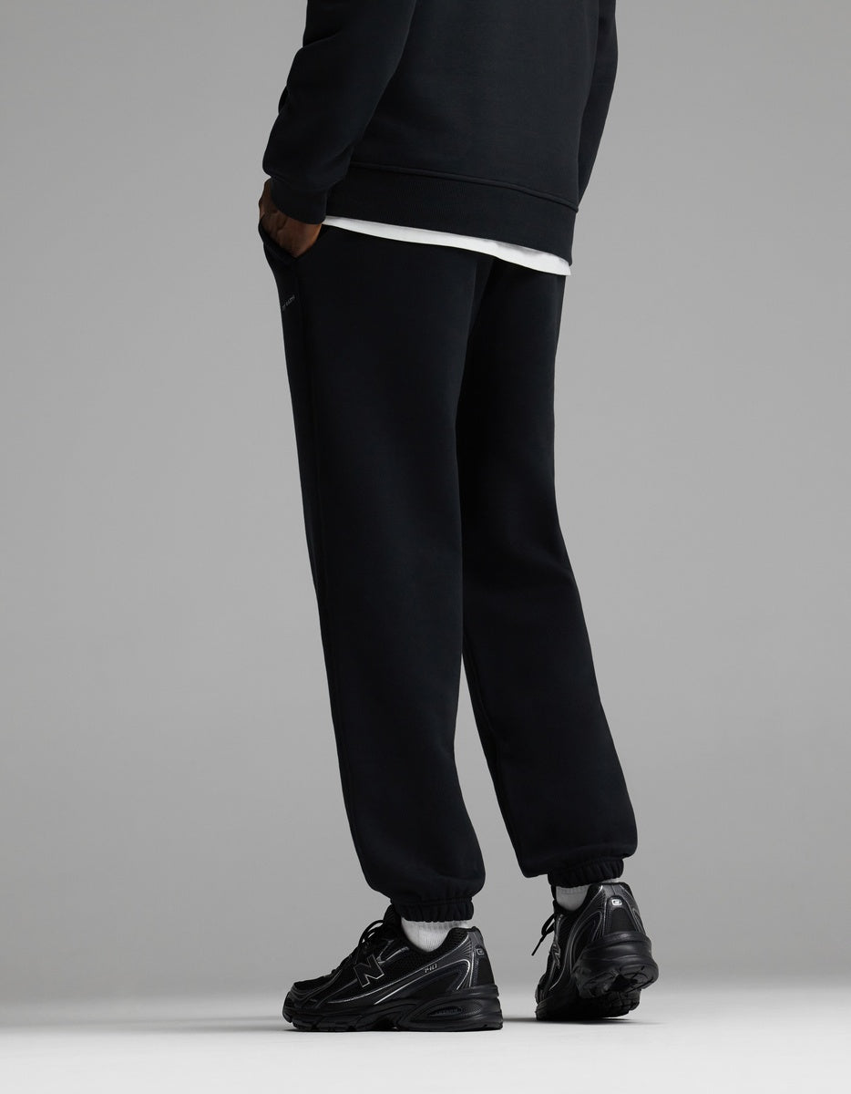 A person wearing Cozy Earth Men’s Cityscape 2.0 Sweatpant in black stands facing away, with only their legs and lower torso visible, against a plain gray background.