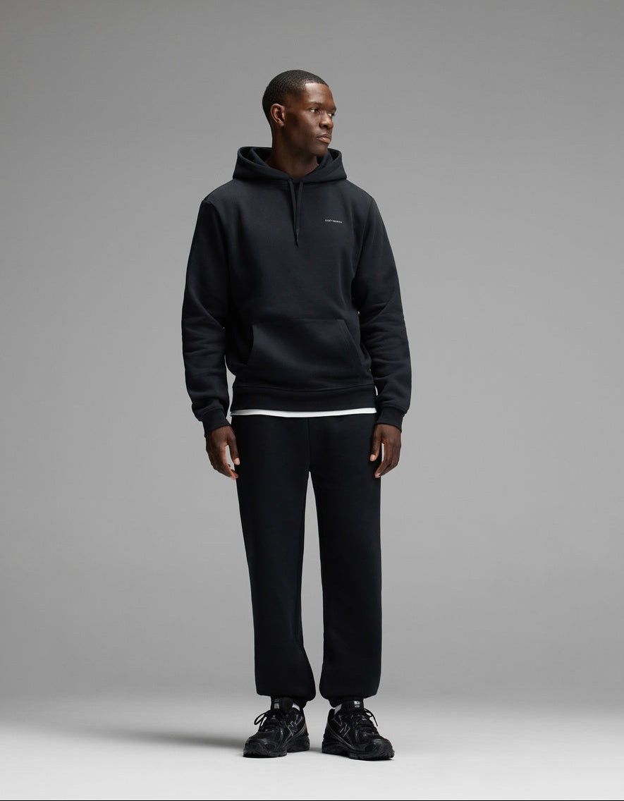 A man wearing Cozy Earth’s Men’s Cityscape 2.0 Sweatpant in black, along with a matching black hoodie and sneakers, stands against a plain gray background, looking to his left.
