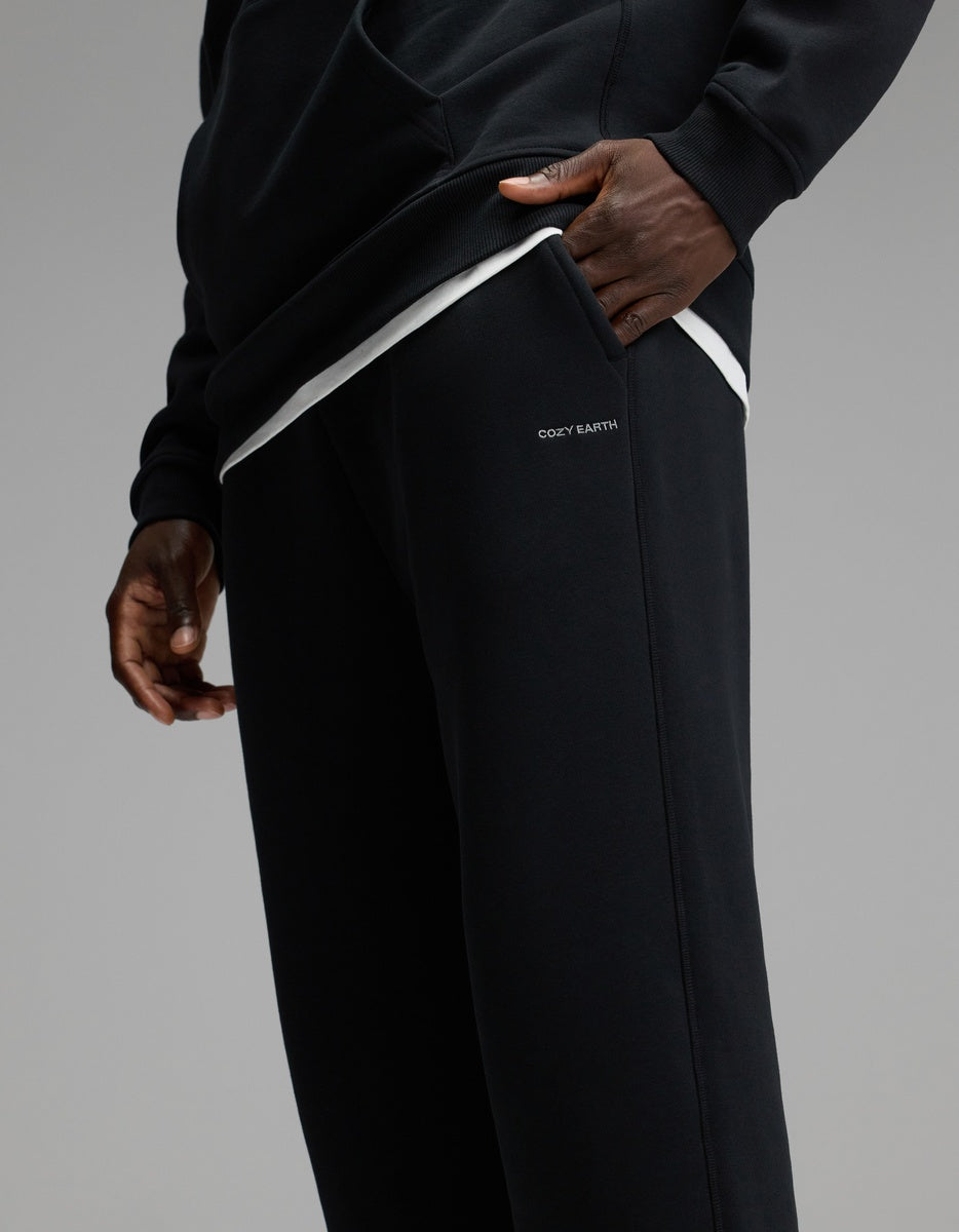 A person wearing the Cozy Earth Men’s Cityscape 2.0 Sweatpant in black, paired with a matching black sweatshirt, stands with one hand in their pocket. The sweatpants feature “COZY EARTH” printed near the pocket, set against a plain gray background.
