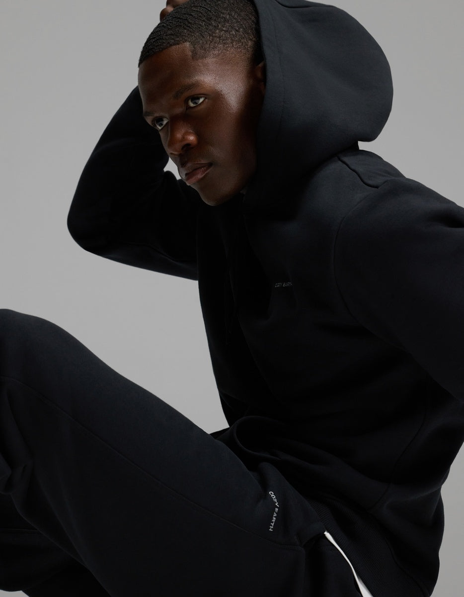 A man wearing Cozy Earth’s Men’s Cityscape 2.0 Sweatpant in black sits in a relaxed pose against a plain gray background, looking to the side with one hand on his head.