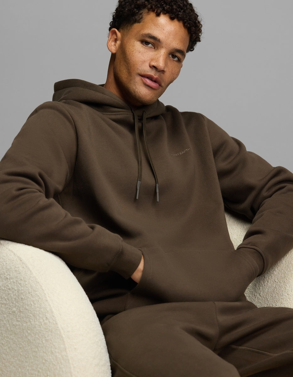 A man wearing a brown Men’s Cityscape Hoodie & Sweatpant Set, sitting in a white textured armchair.