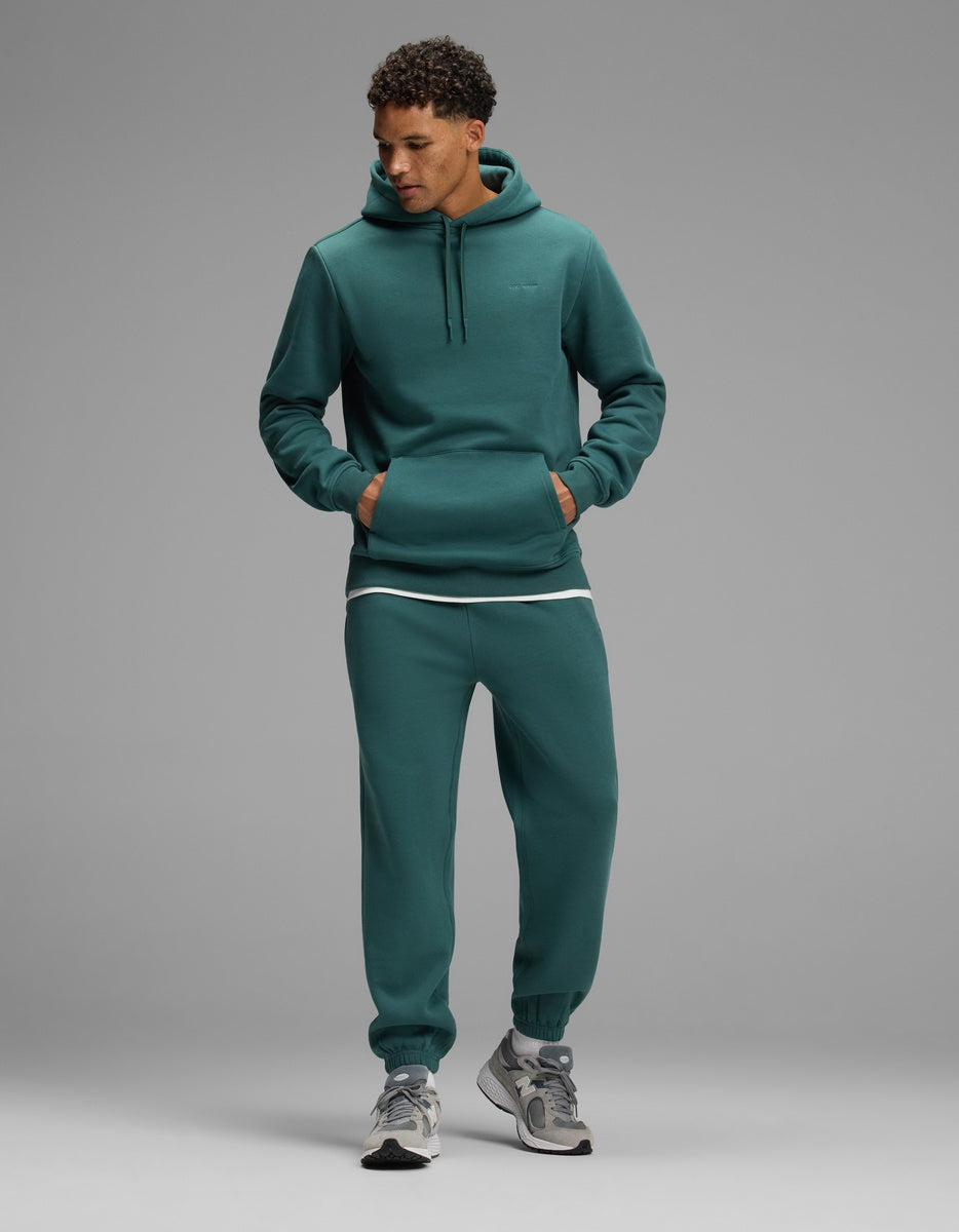 A man wearing a teal Men’s Cityscape Hoodie & Sweatpant Set with a white undershirt and grey sneakers.