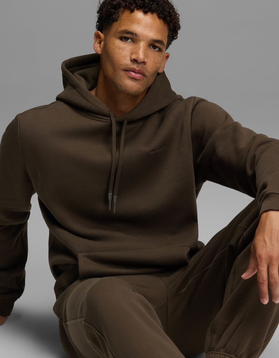 A man wears a dark brown long-sleeved Men’s Cityscape Hoodie with a front pocket and matching sweatpants.