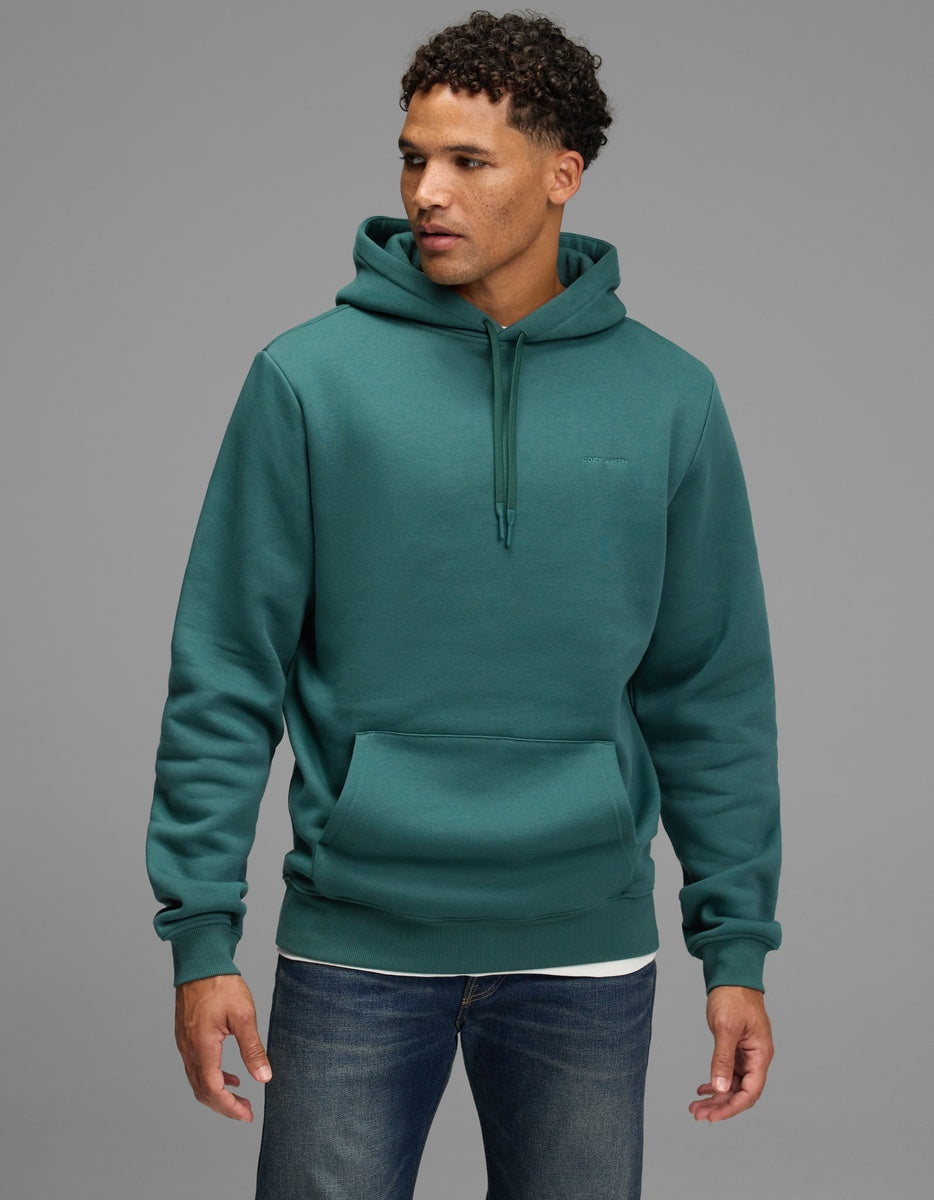 A man wearing a dark teal Cozy Earth Men’s Cityscape Hoodie with a drawstring hood and a front kangaroo pocket.