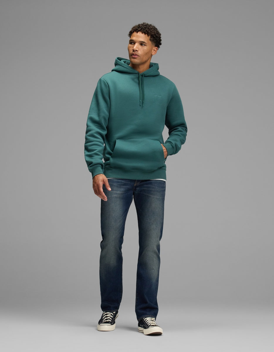 A man wearing a dark teal Cozy Earth Men’s Cityscape Hoodie with a front pouch pocket, blue jeans, and black sneakers.