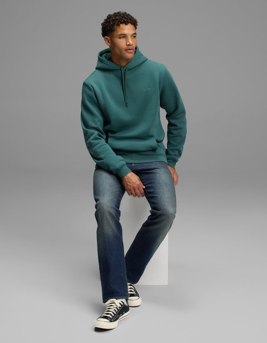 A man wearing a teal Cozy Earth Men’s Cityscape Hoodie, blue jeans, and black low-top sneakers sits on a white block.