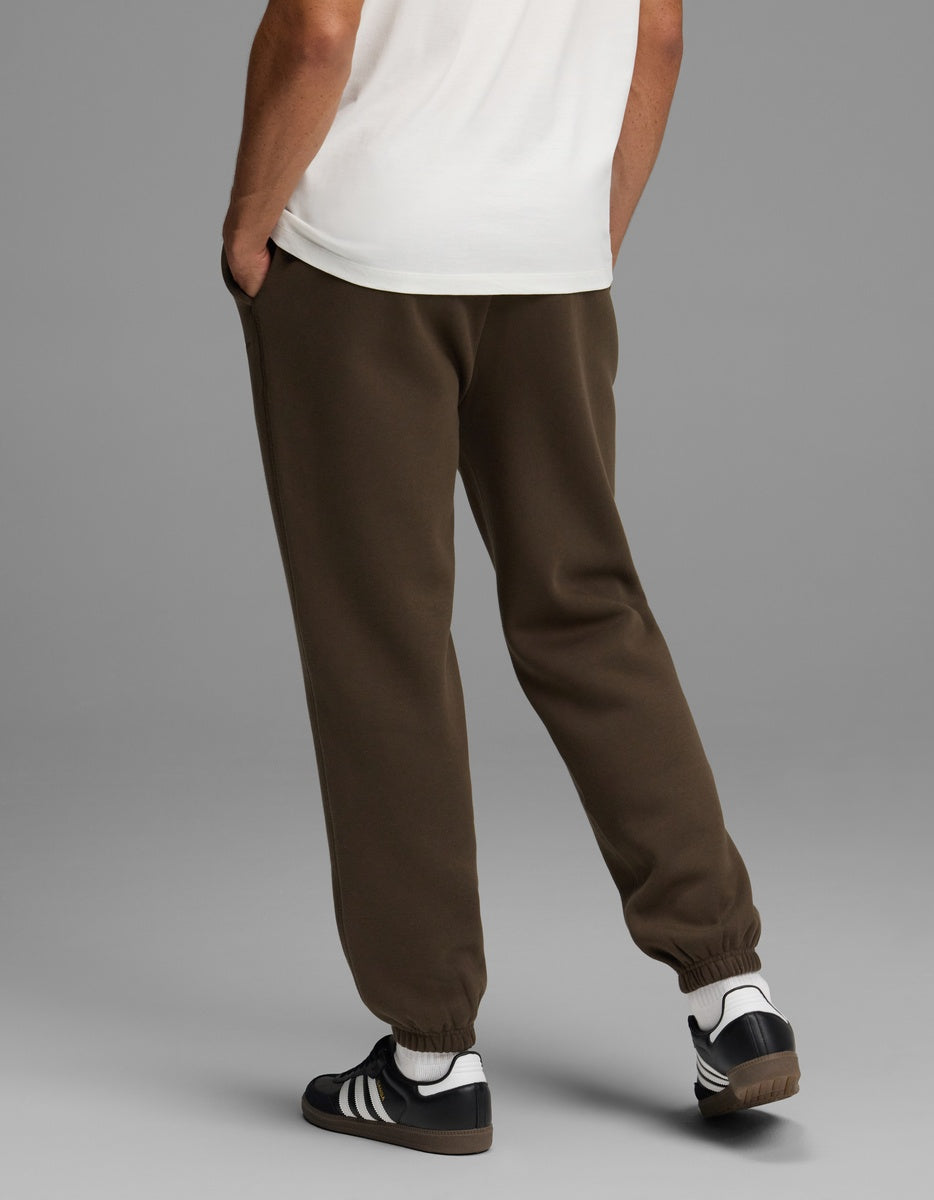 Back view of a person wearing dark brown Men’s Cityscape Sweatpant with elastic cuffs, a white t-shirt, and black and white sneakers.
