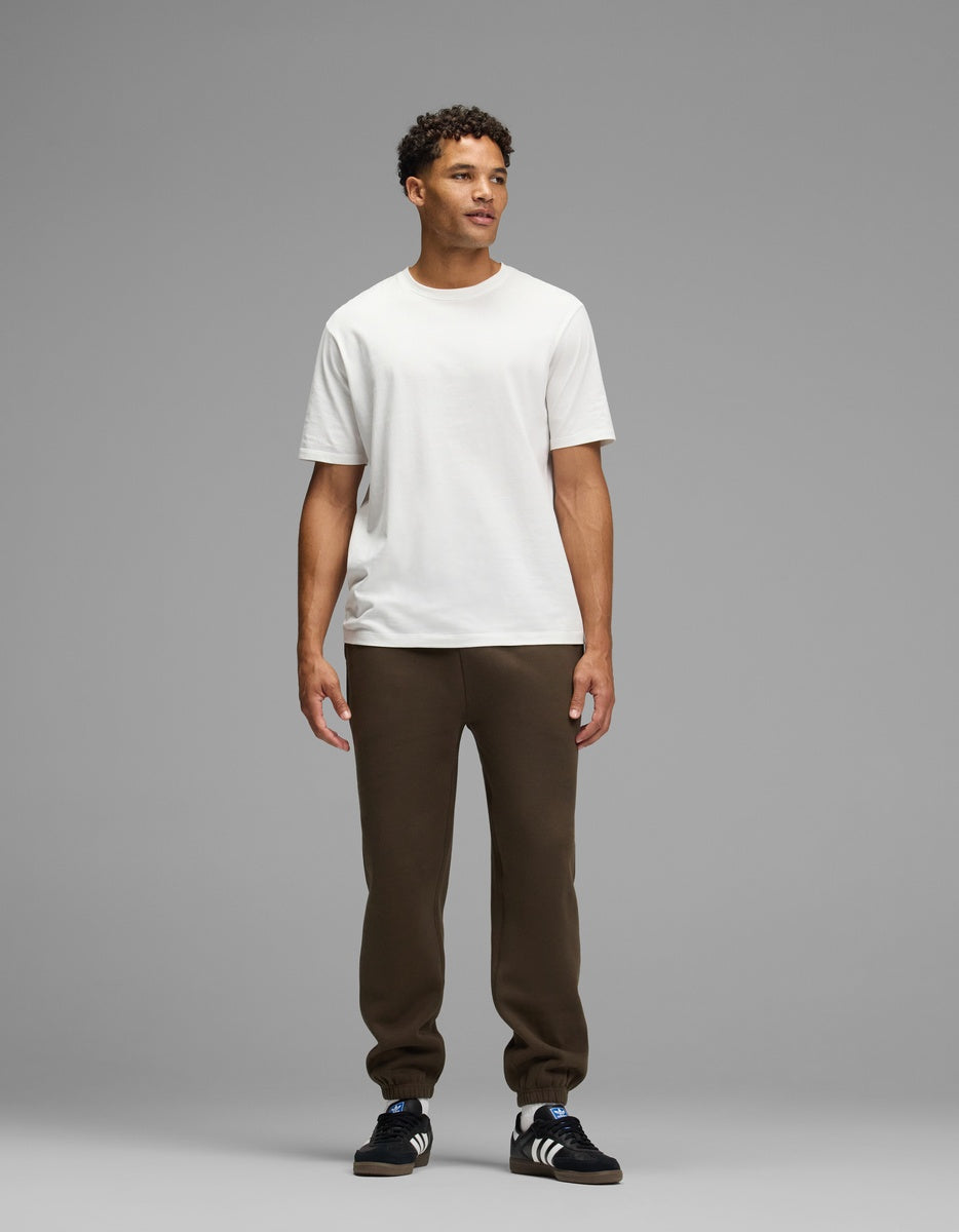A man wearing a white t-shirt, Men’s Cityscape Sweatpant in dark brown with ribbed cuffs, and black sneakers.