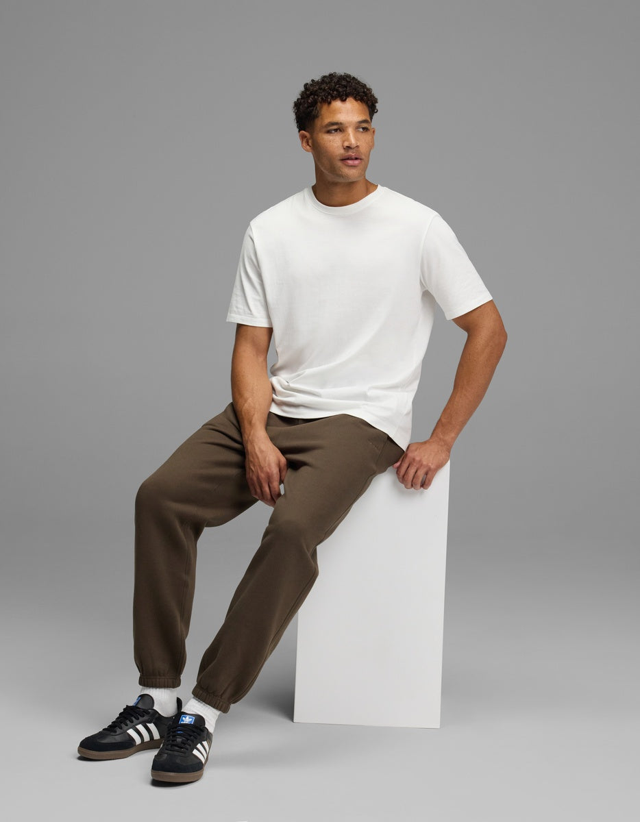 A man wearing a white t-shirt and brown Men’s Cityscape Sweatpant sits on a white block.