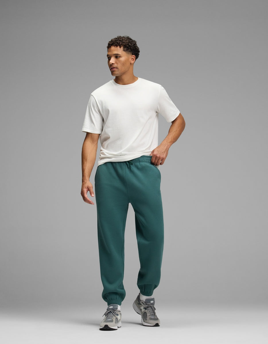A man wearing a white t-shirt and teal Men’s Cityscape Sweatpant with elastic cuffs, paired with gray sneakers.
