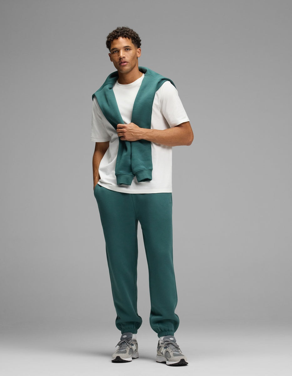 A man wears a white t-shirt, matching dark teal Men’s Cityscape Sweatpant, and a dark teal sweatshirt draped over his shoulders.