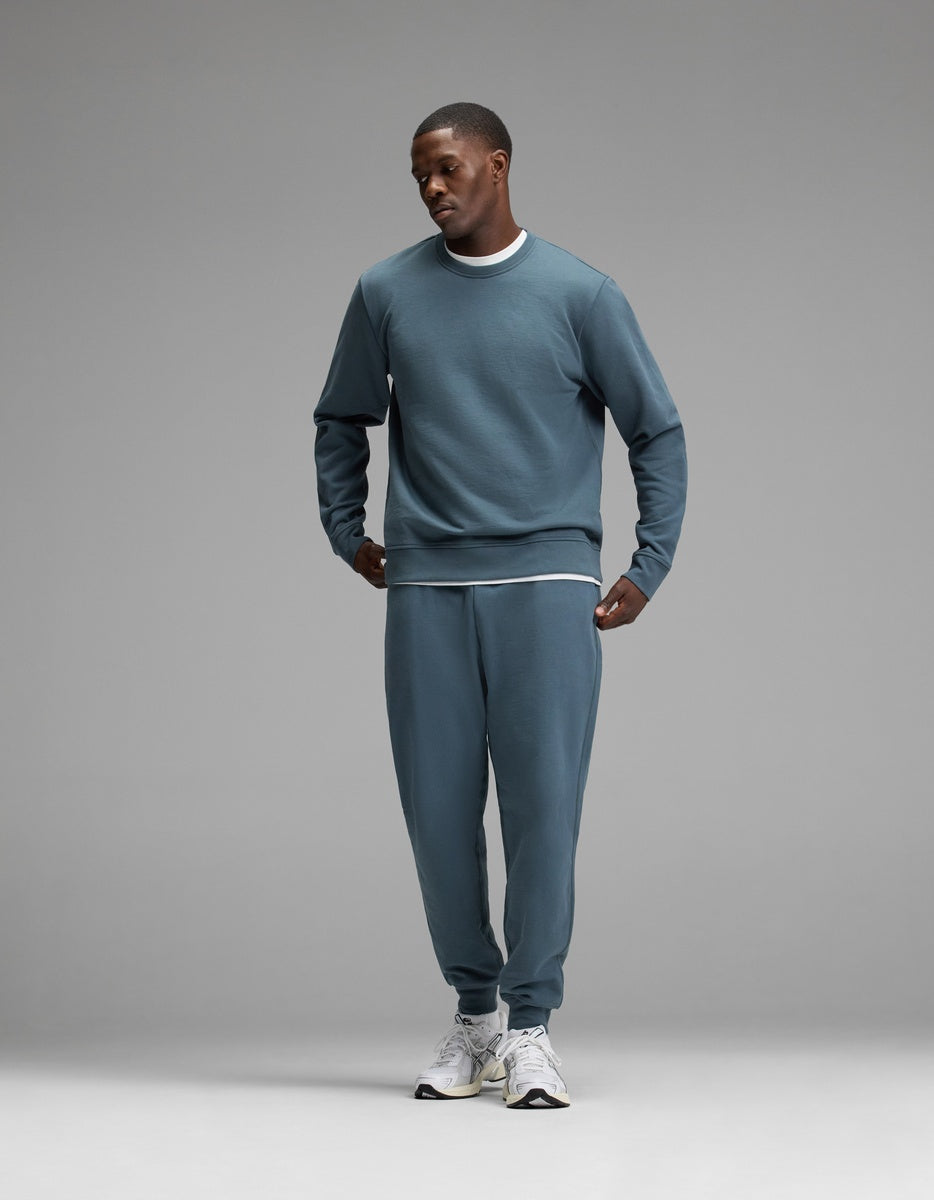 A man wearing the Cozy Earth Men’s Dylan Crewneck & Jogger Set in blue, paired with white sneakers, stands against a plain gray background, looking down with one hand in his pocket.