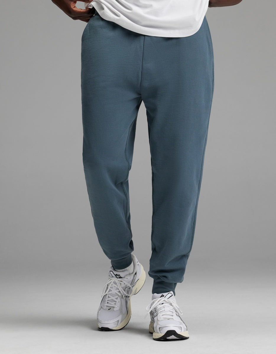 A person is shown from the waist down wearing Cozy Earth’s Men’s Dylan Jogger in blue, paired with a white shirt and white athletic sneakers, standing against a plain gray background.