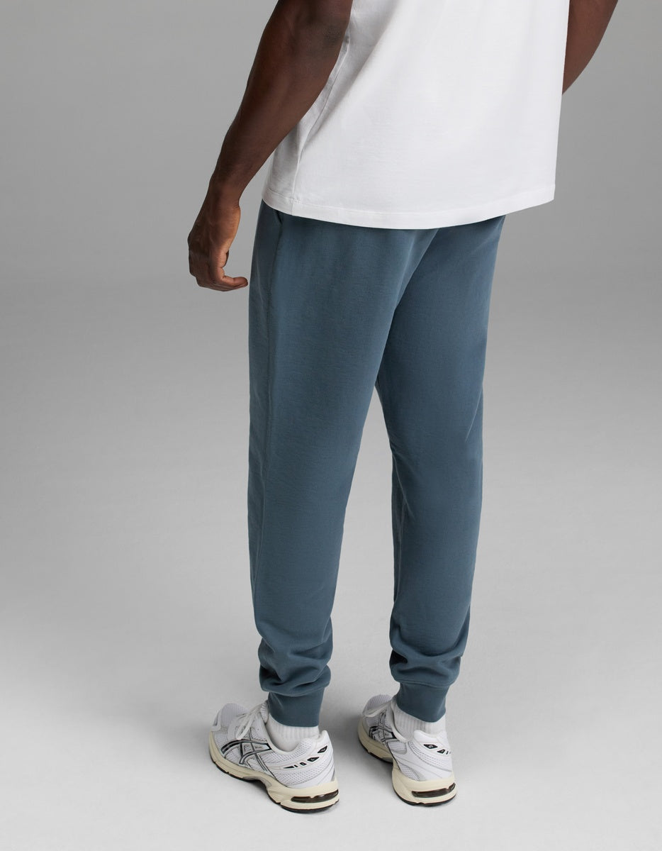 A person wearing a white T-shirt, Cozy Earth Men’s Dylan Jogger in blue, and white sneakers stands facing away from the camera on a gray background.