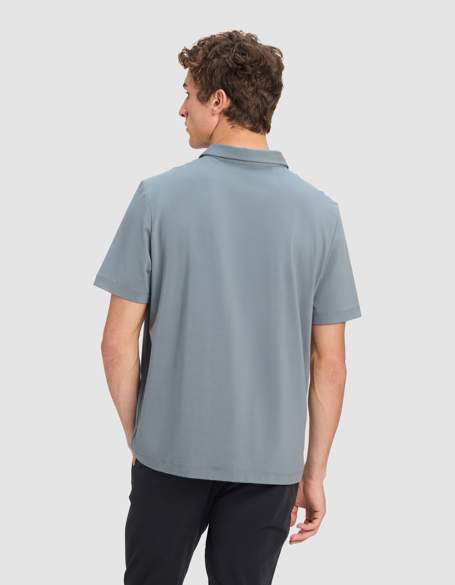 A man with short curly hair is seen from behind wearing the Cozy Earth Men's Everyday Polo in light blue with black pants, set against a plain light gray background.