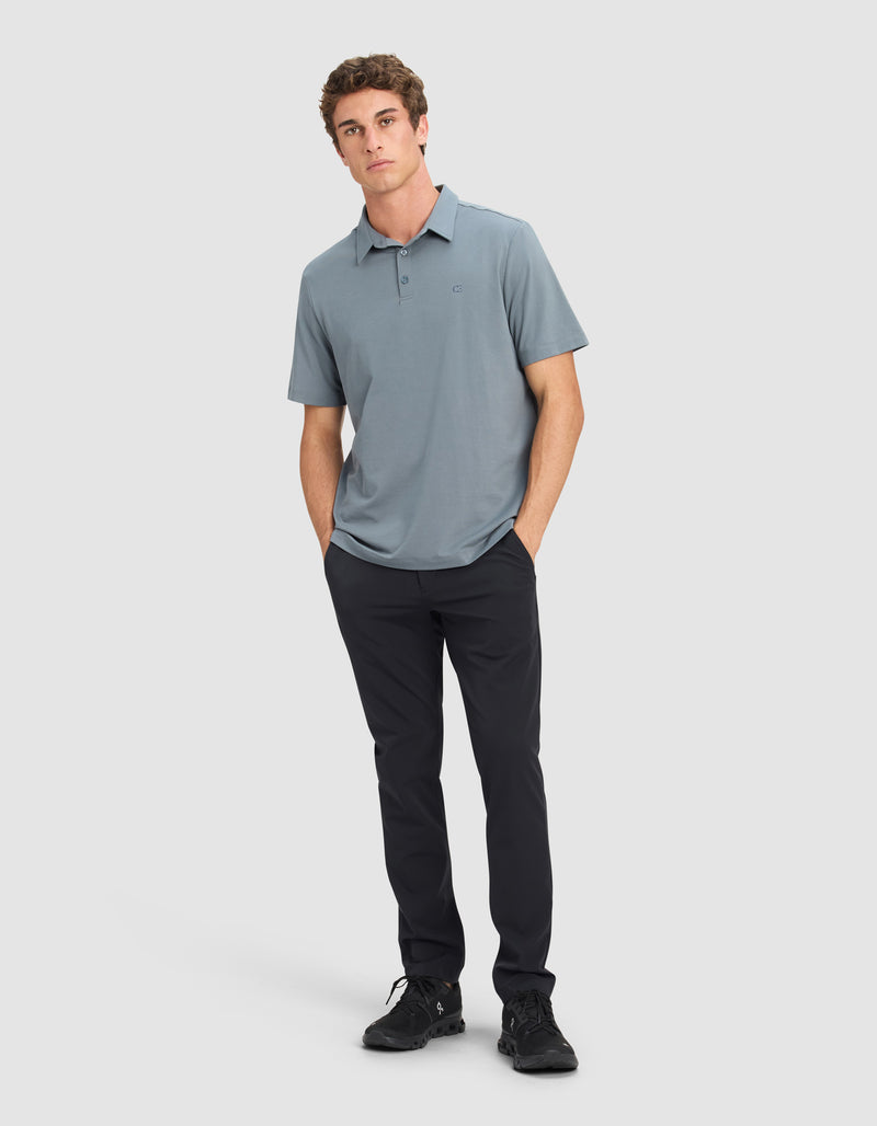 A man wearing the Cozy Earth Men's Everyday Polo in light blue, paired with dark pants and black sneakers, stands with his hands in his pockets against a plain light gray background.
