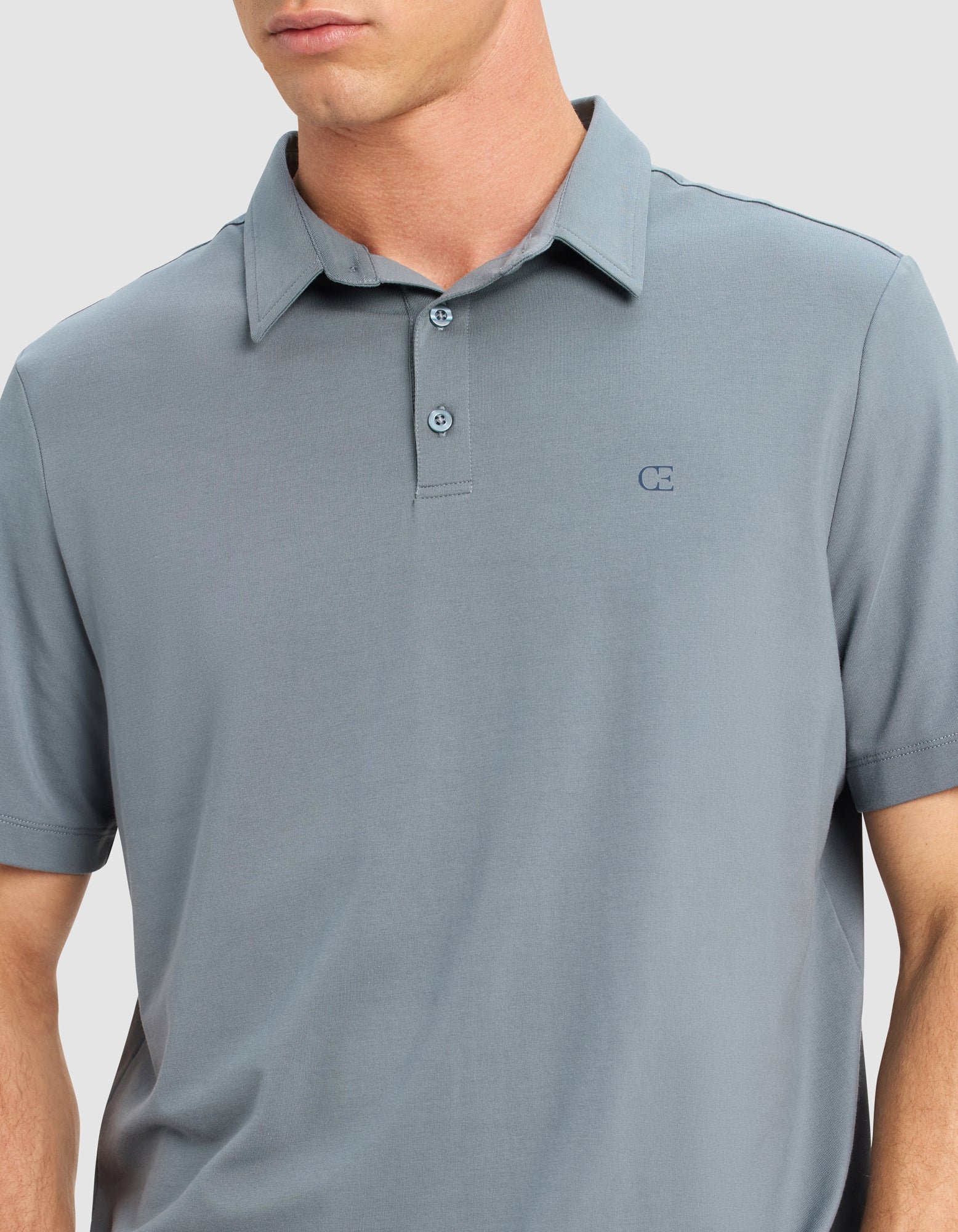 A man models the Cozy Earth Men's Everyday Polo in light gray, featuring short sleeves and a small embroidered logo on the left chest, standing against a plain light background. The image is cropped from his lips to his waist.