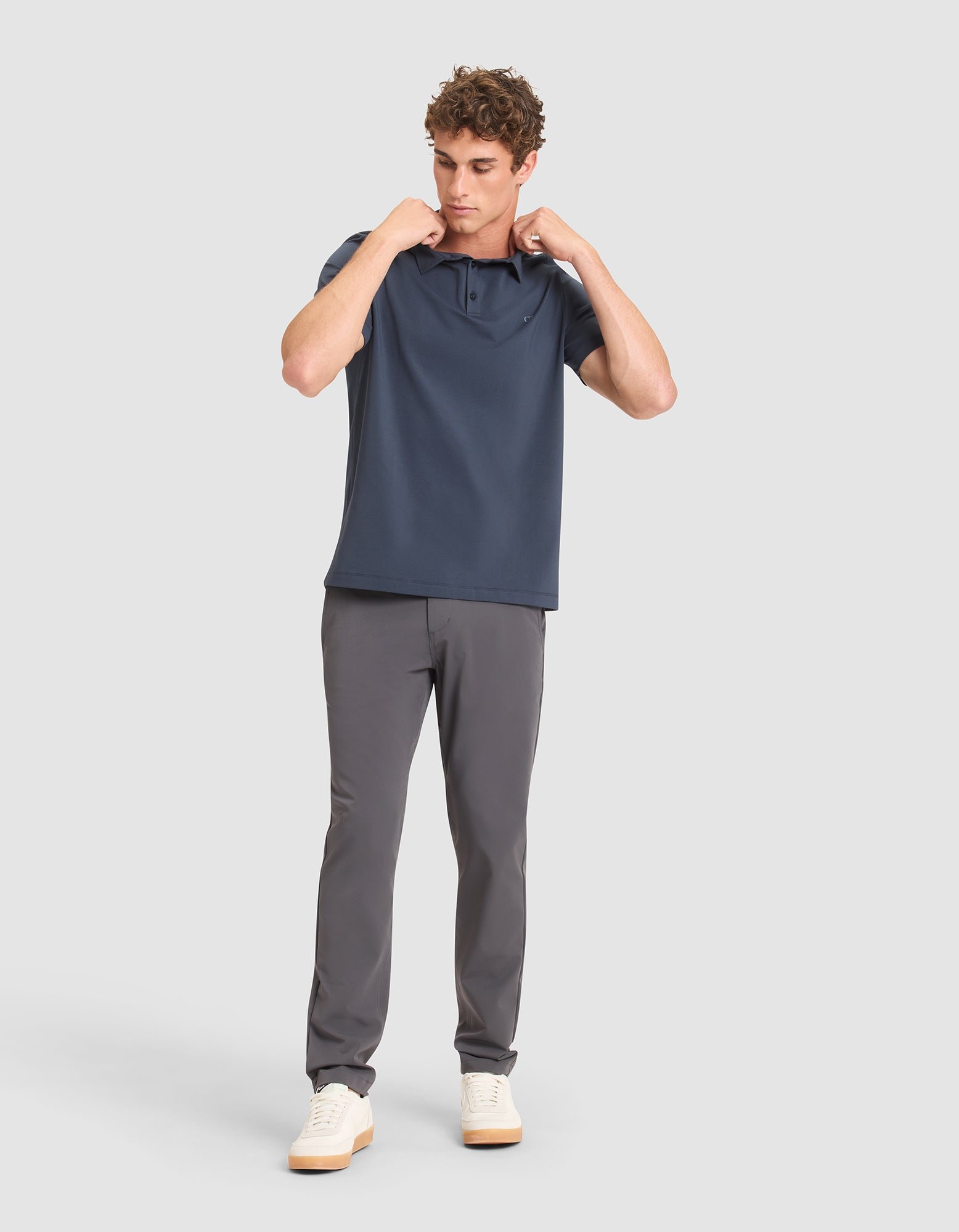 A man stands against a light gray background, wearing Cozy Earth's Men's Everyday Polo in navy with gray pants and white sneakers. He looks down while adjusting the collar of his shirt with both hands. 