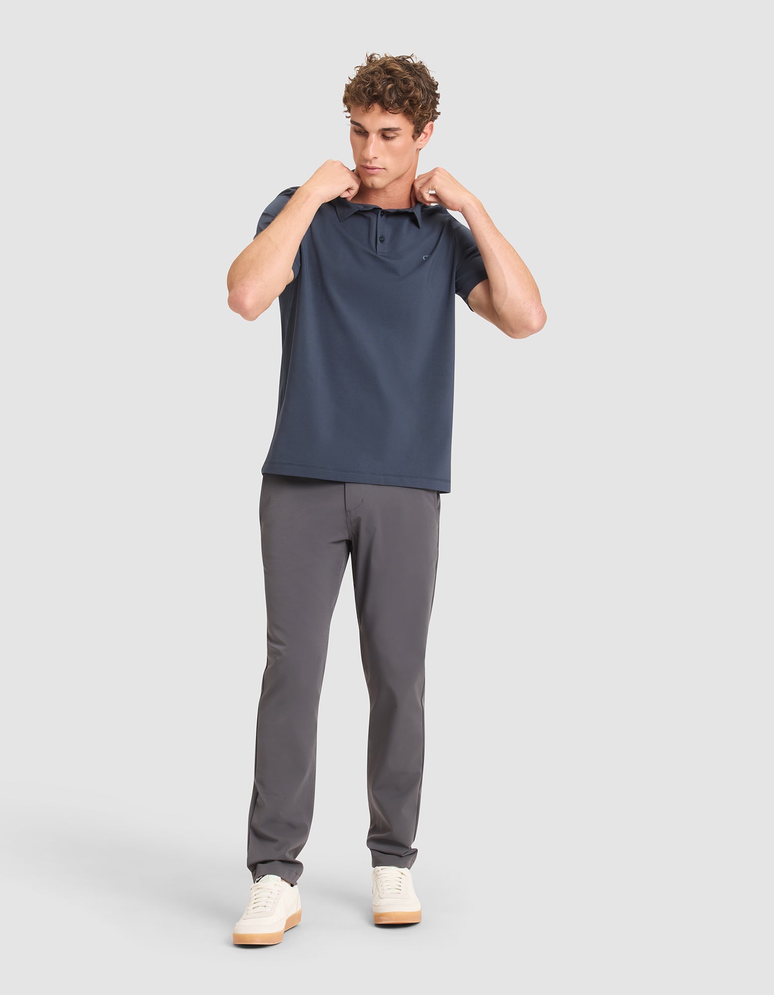 A man stands against a light gray background, wearing Cozy Earth's Men's Everyday Polo in navy with gray pants and white sneakers. He looks down while adjusting the collar of his shirt with both hands. 