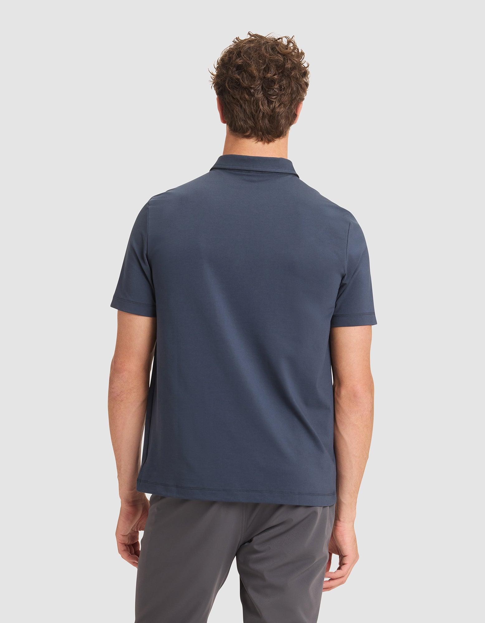 A man with short, curly brown hair is seen from behind wearing Cozy Earth's Men's Everyday Polo in navy blue and gray pants, standing against a plain light background. 