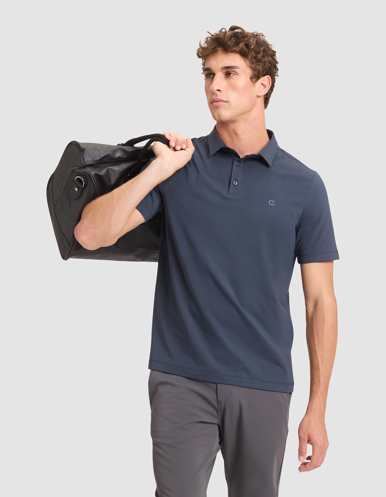 A man wears the Cozy Earth Men's Everyday Polo in blue with gray pants and carries a black duffel bag over his shoulder, posed against a plain light gray background. 
