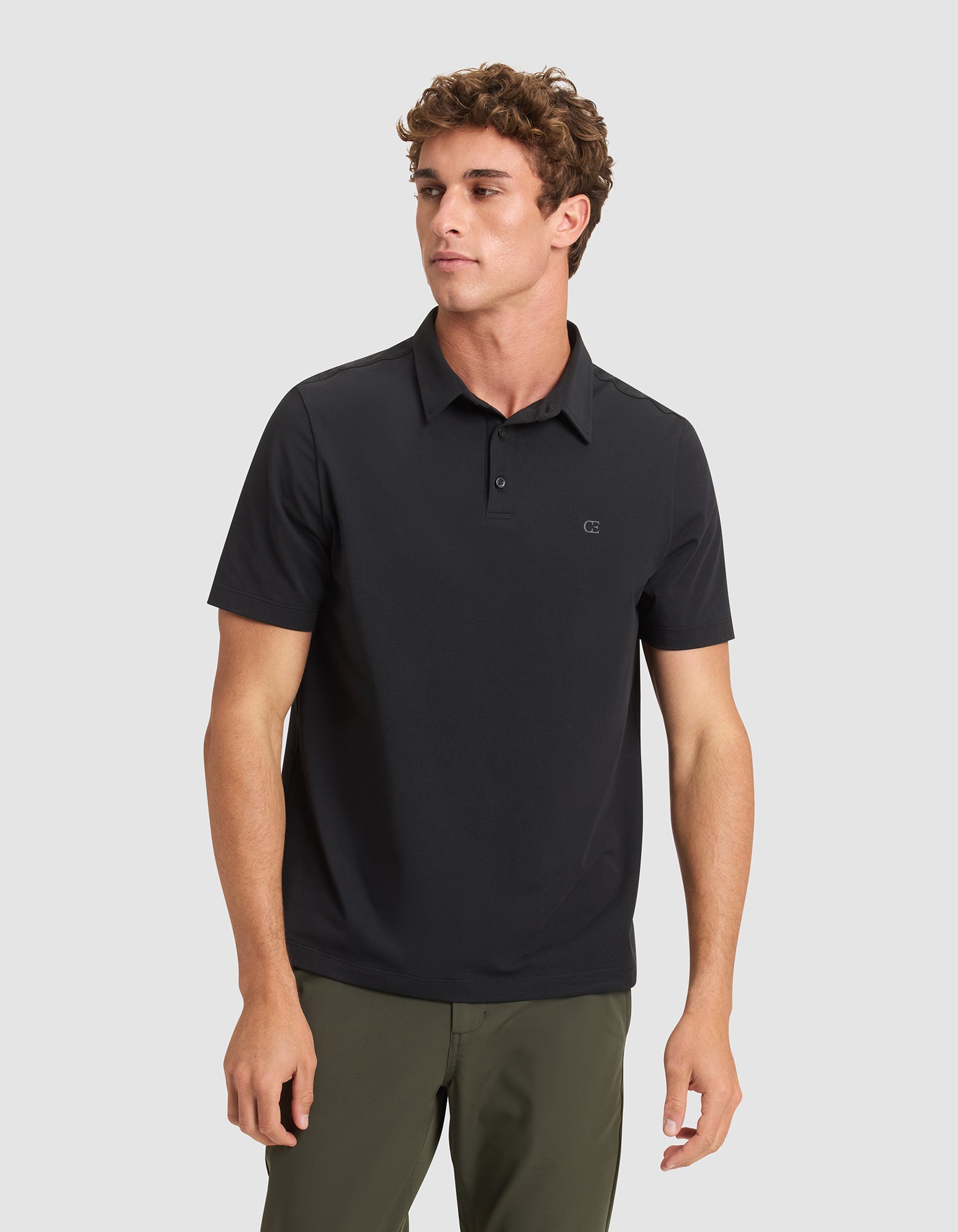 A young man with short curly hair models the Cozy Earth Men's Everyday Polo in black, paired with olive green pants, standing against a light gray background and gazing slightly to his left. |Color:Jet Black
