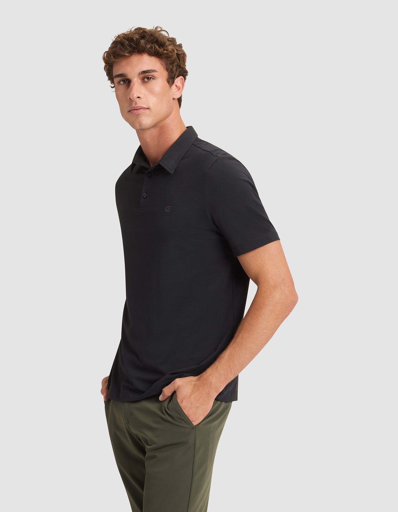 A man stands with his hands in his pockets, wearing the Cozy Earth Men's Everyday Polo in black and olive green pants, looking slightly to the side against a plain light gray background. 