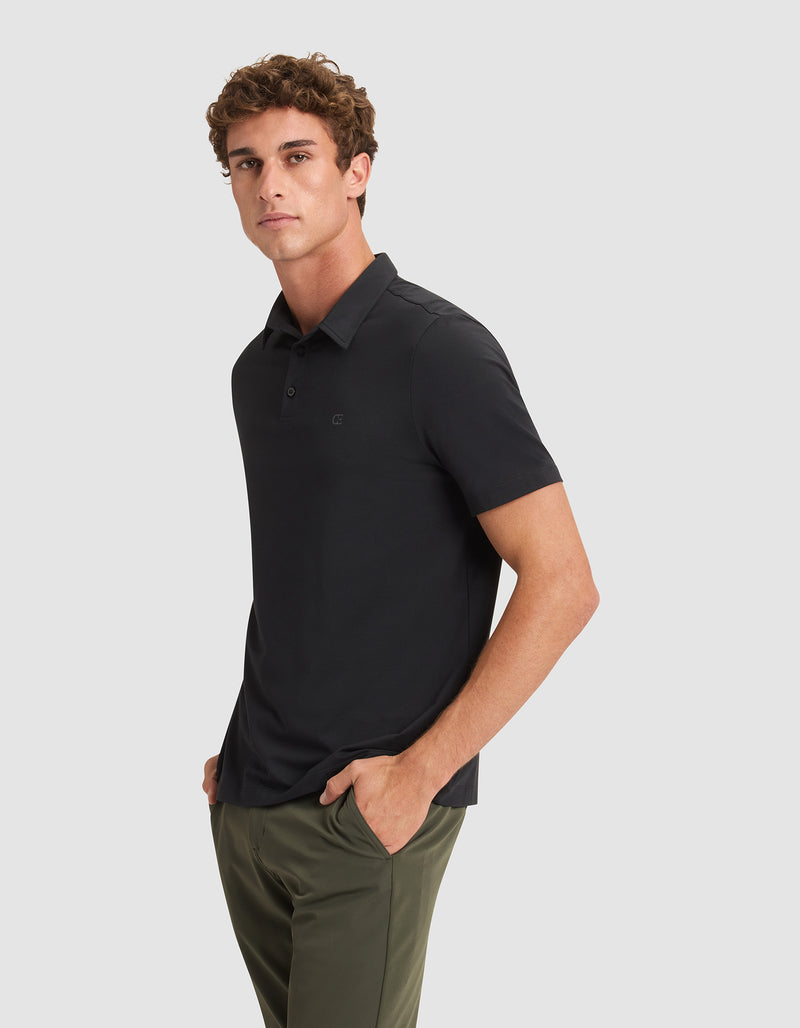 A man stands with his hands in his pockets, wearing the Cozy Earth Men's Everyday Polo in black and olive green pants, looking slightly to the side against a plain light gray background.