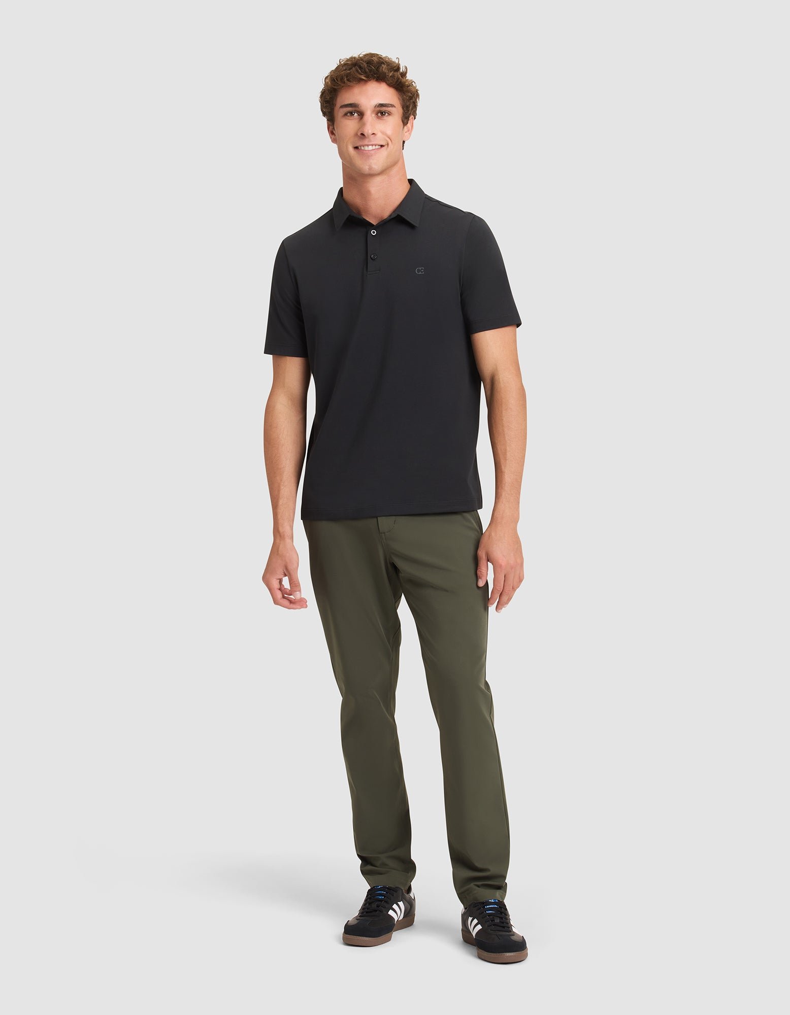 A man stands against a light gray background, smiling and facing forward with one hand by his side. He wears the Cozy Earth Men's Everyday Polo in black, paired with olive green pants and black sneakers with white accents. 