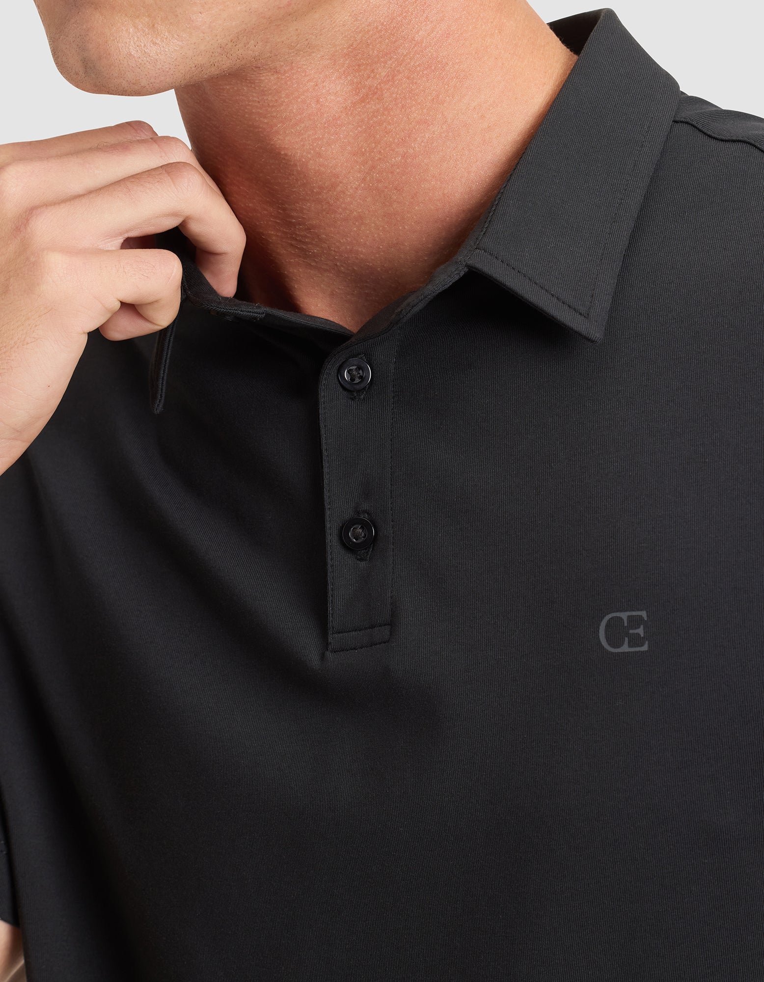 A man wears the Cozy Earth Men's Everyday Polo in black, featuring a two-button collar and a subtle "CE" logo on the chest. His hand is near his neck, and his face is partially visible. 