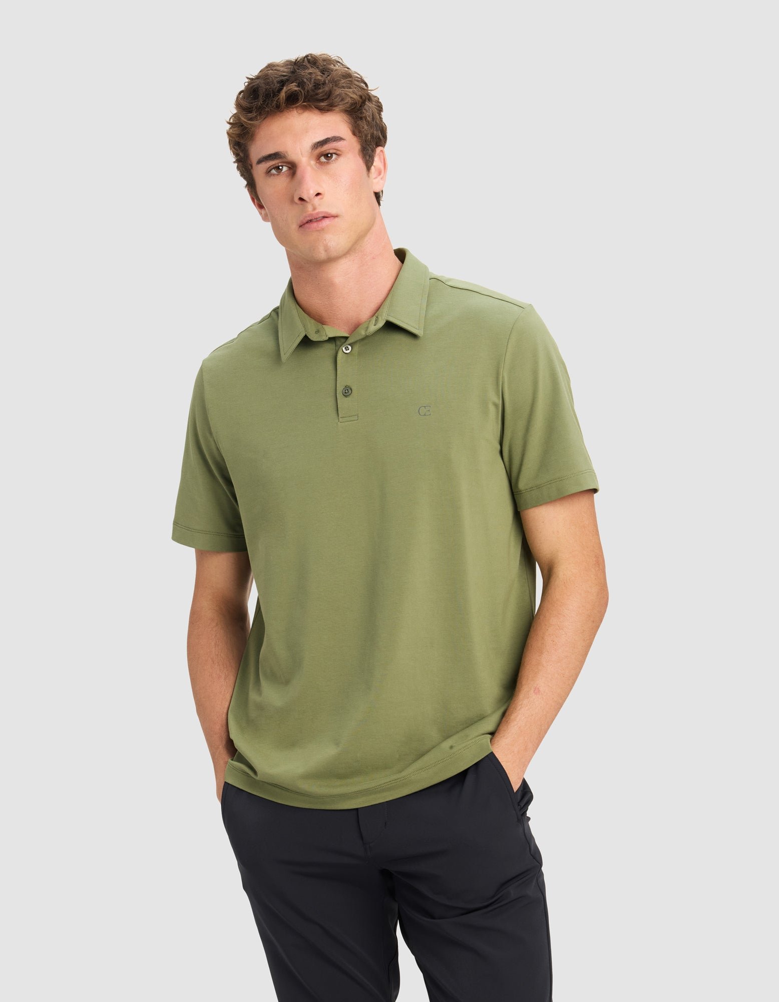 A man models the Cozy Earth Men's Everyday Polo in light green with black pants, standing hands-in-pockets against a plain light gray background.