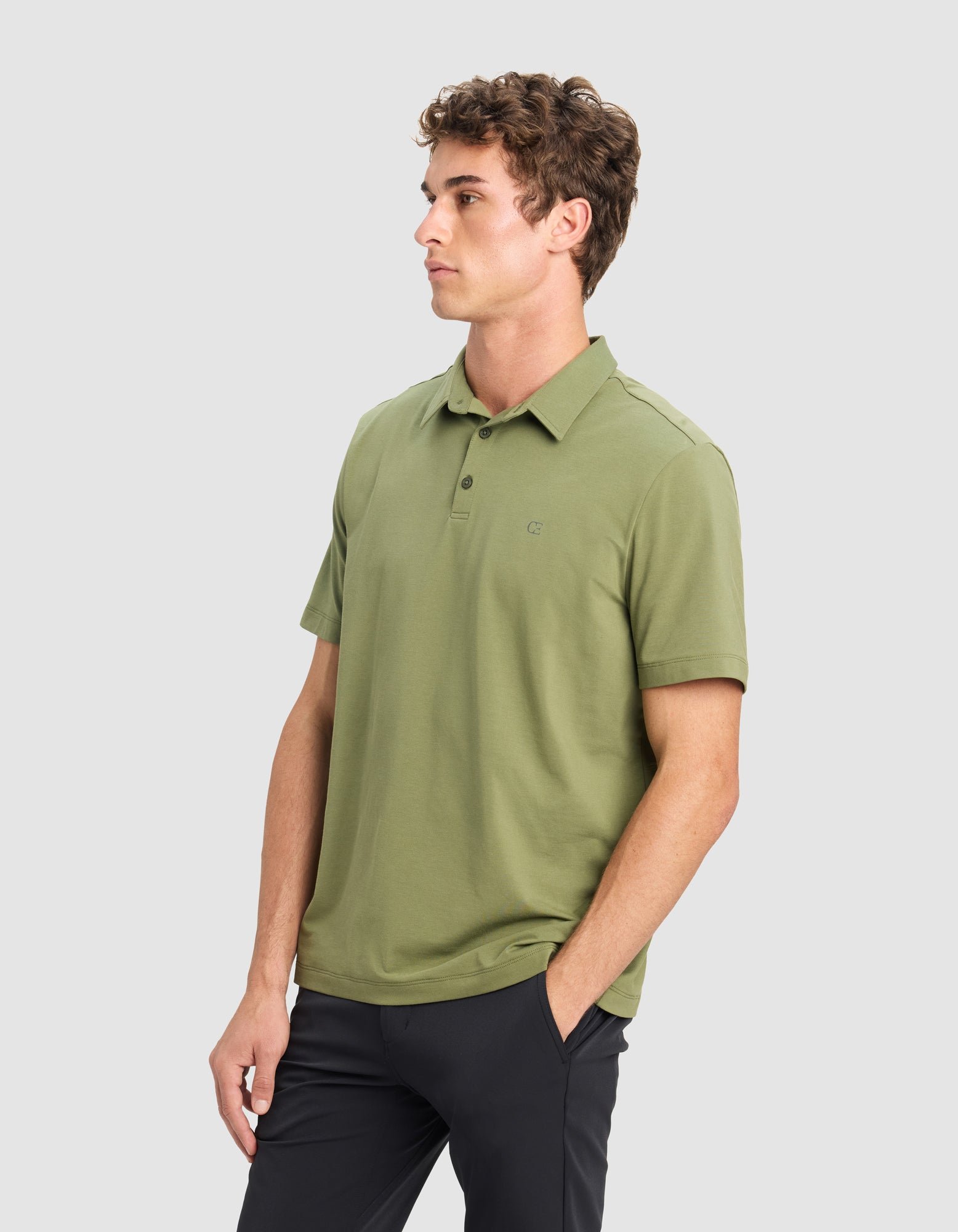 A man with short curly hair models the Cozy Earth Men's Everyday Polo in light olive green with dark pants, standing with one hand in his pocket and looking to the side against a plain light gray background. 