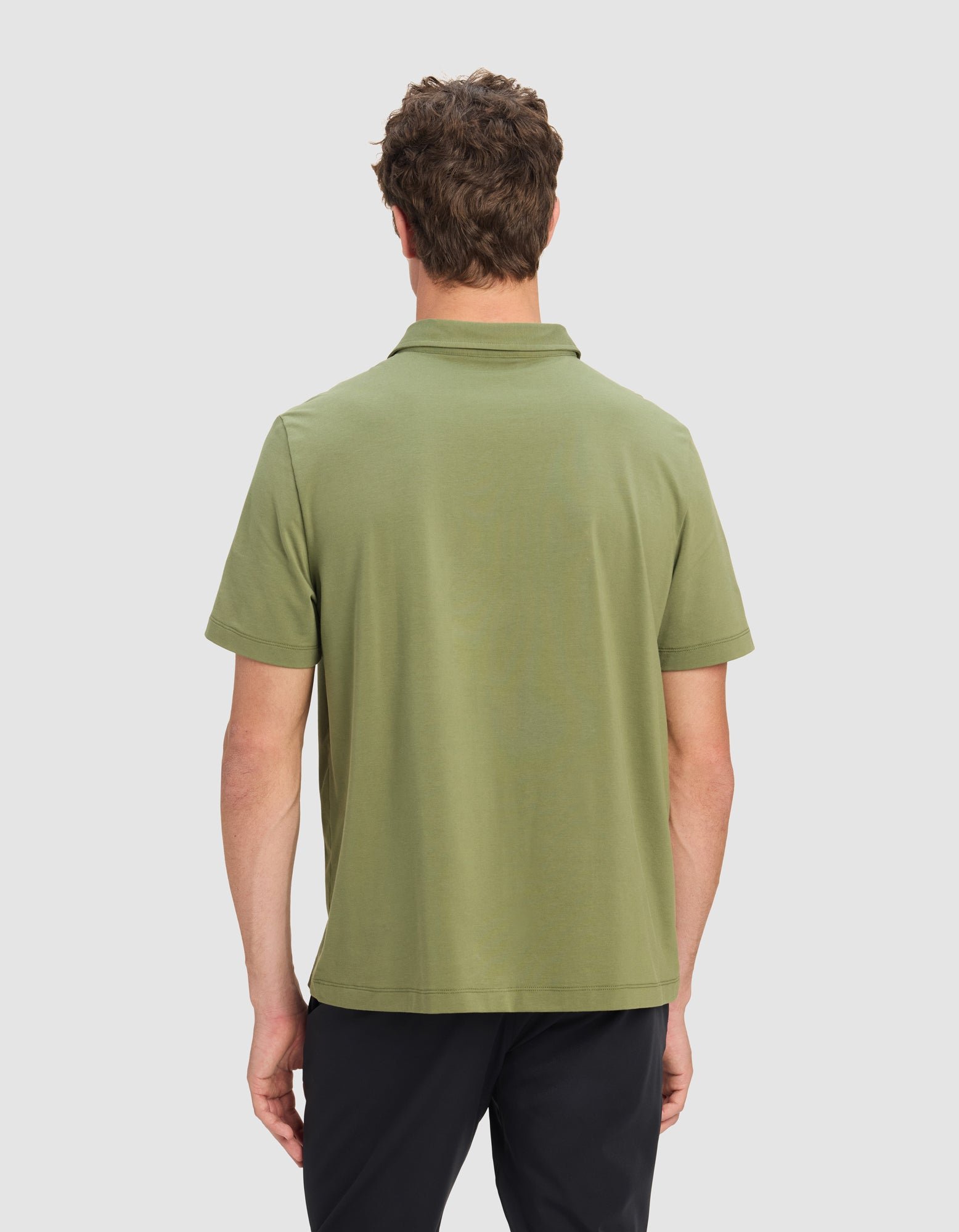 A man is seen from the back, wearing Cozy Earth's Men's Everyday Polo in light olive green and dark pants, set against a plain light gray background.