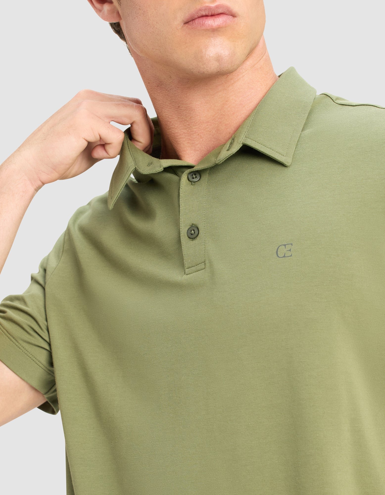 A man adjusts the collar of a Cozy Earth Men's Everyday Polo in light olive green, featuring a small chest logo, button placket, and short sleeves. The background is light gray.