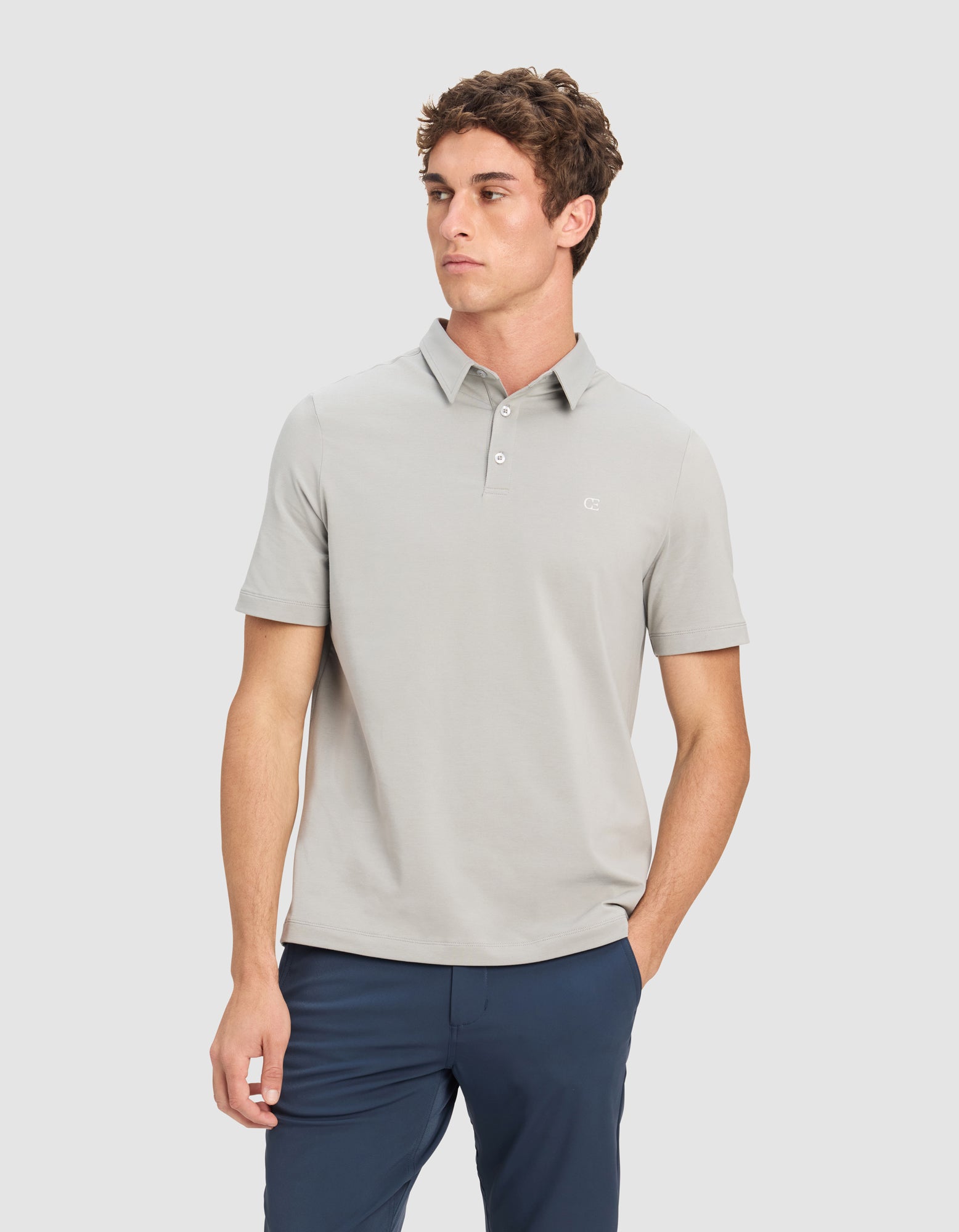 A man models the Cozy Earth Men's Everyday Polo in light gray, paired with dark blue pants, standing with one hand in his pocket against a plain light background.