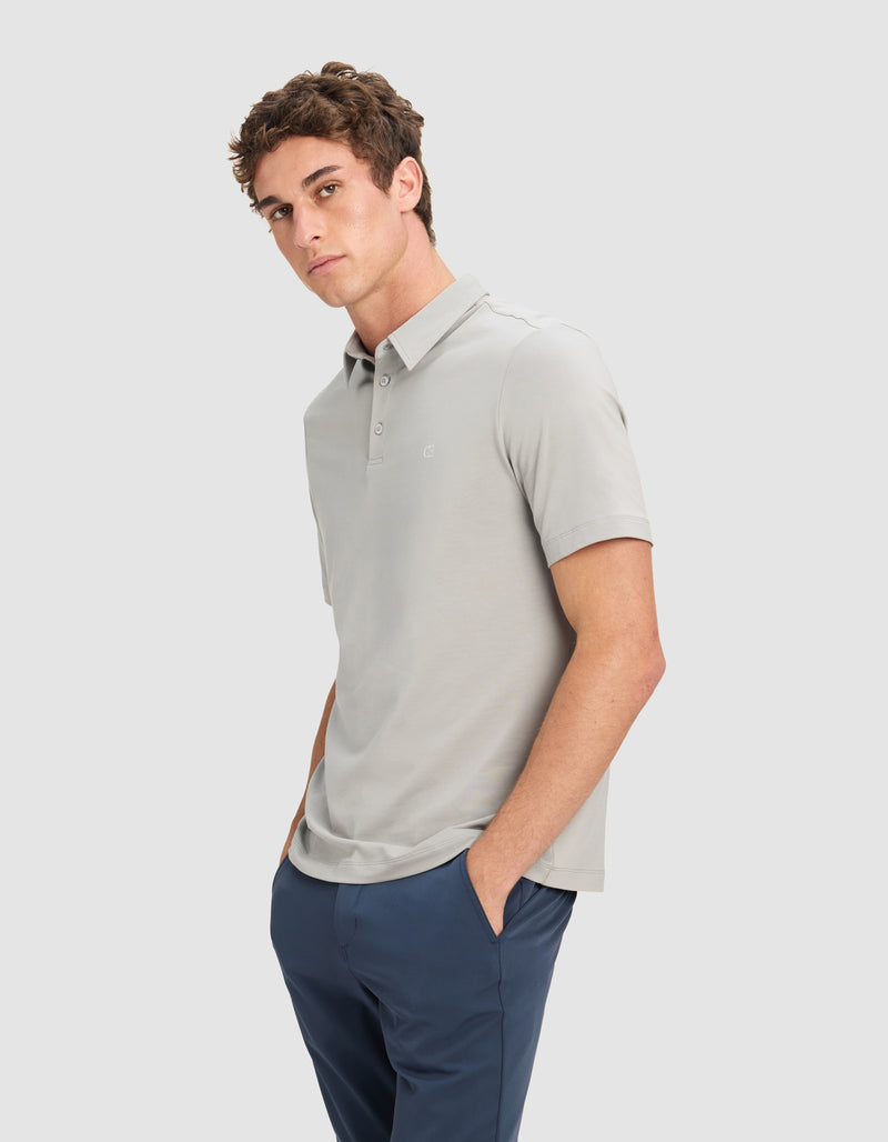 A man with short, curly brown hair wears the Cozy Earth Men's Everyday Polo in light gray and navy pants. He stands against a plain light background, hands in his pockets, gazing slightly to the side.