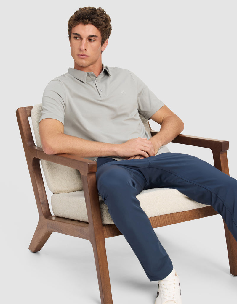 A man wearing the Cozy Earth Men's Everyday Polo sits on a cushioned wooden chair, paired with navy pants and white sneakers, looking off to the side against a plain background.