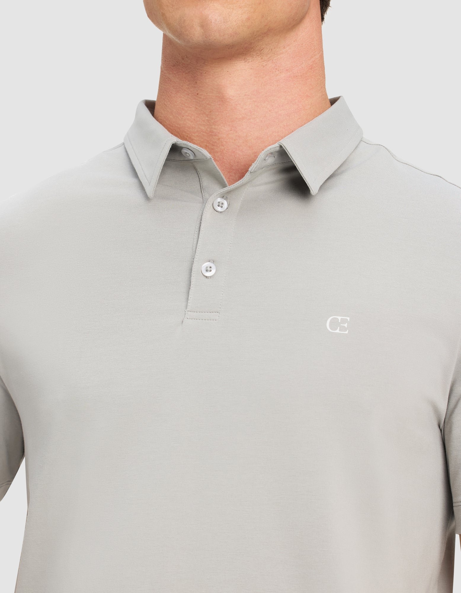 A person wears the Cozy Earth Men's Everyday Polo in light gray, featuring a three-button collar and a small white "CE" logo on the left chest. The image is cropped to show only the shirt and neck.