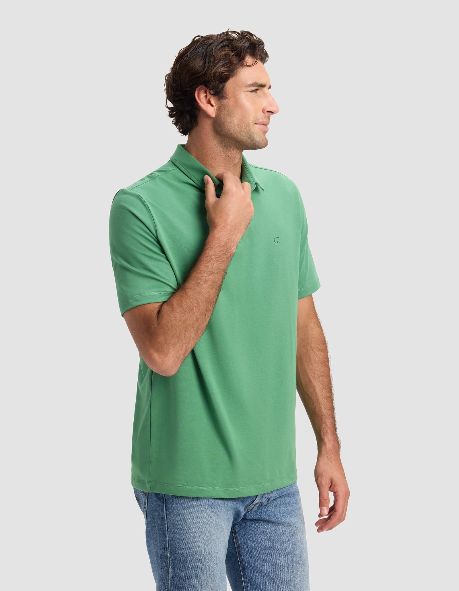 A man with wavy brown hair wears the Cozy Earth Men's Everyday Polo in green and blue jeans, standing sideways with one hand on the collar against a plain light gray background.