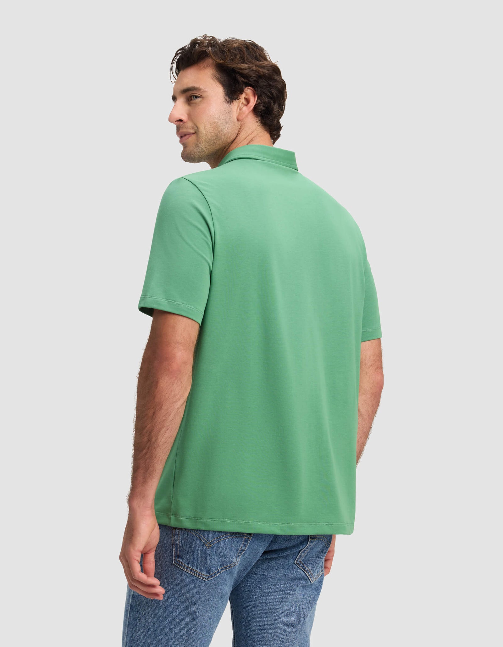 A man stands with his back facing us, turning his head left, wearing the Cozy Earth Men's Everyday Polo (short-sleeve, green) and blue jeans against a plain light gray background.