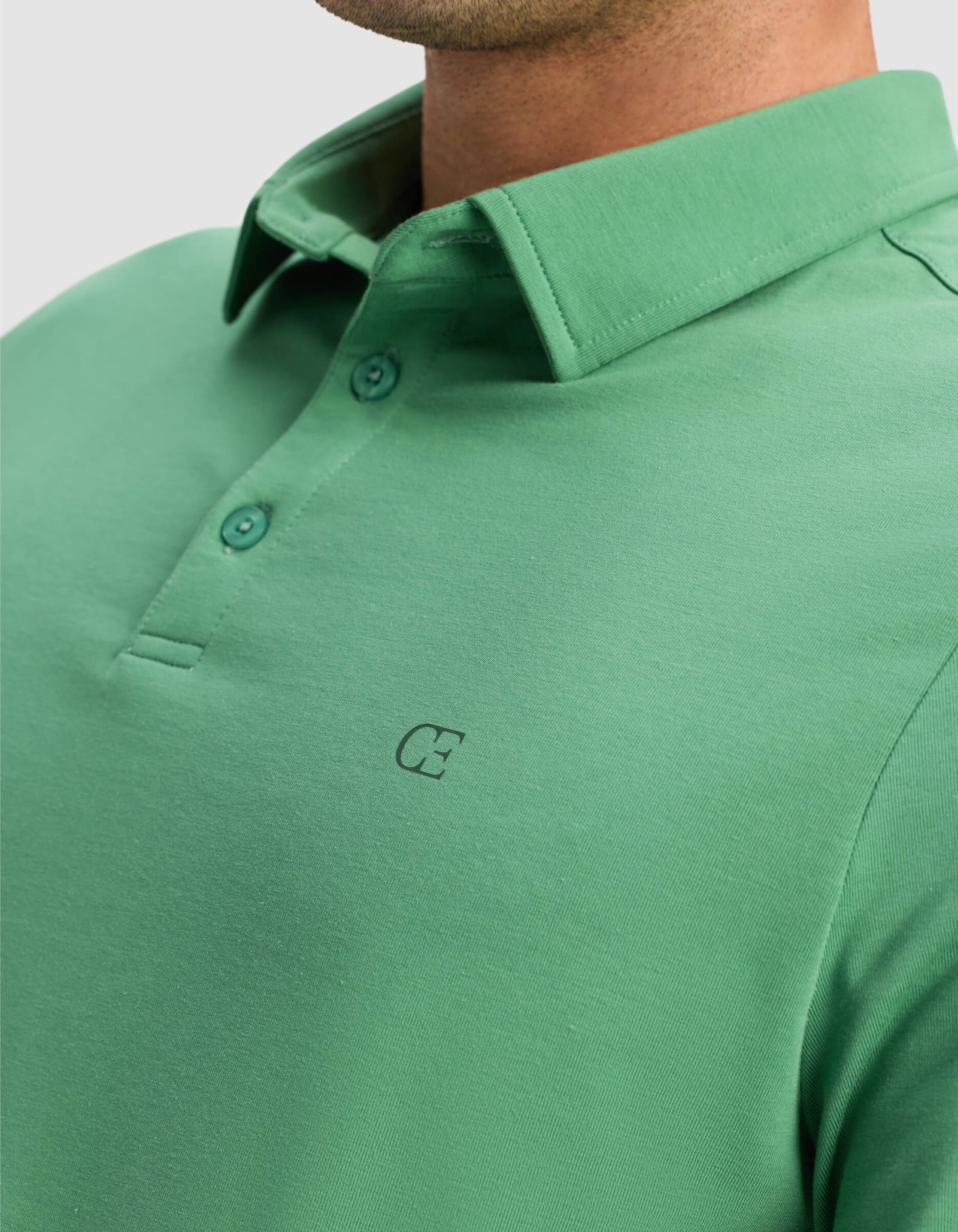 Close-up of a man wearing the Cozy Earth Men's Everyday Polo, featuring a green color, three-button collar, and small "C" chest logo. Only his lower face, neck, and upper torso are visible.