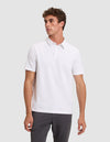 A man with short curly hair wears the Cozy Earth Men's Everyday Polo in plain white and dark pants, standing against a light gray background and looking to his left with a neutral expression. |Color:White