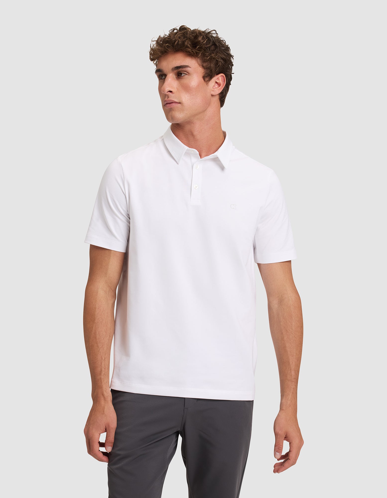 A man with short curly hair wears the Cozy Earth Men's Everyday Polo in plain white and dark pants, standing against a light gray background and looking to his left with a neutral expression. 
