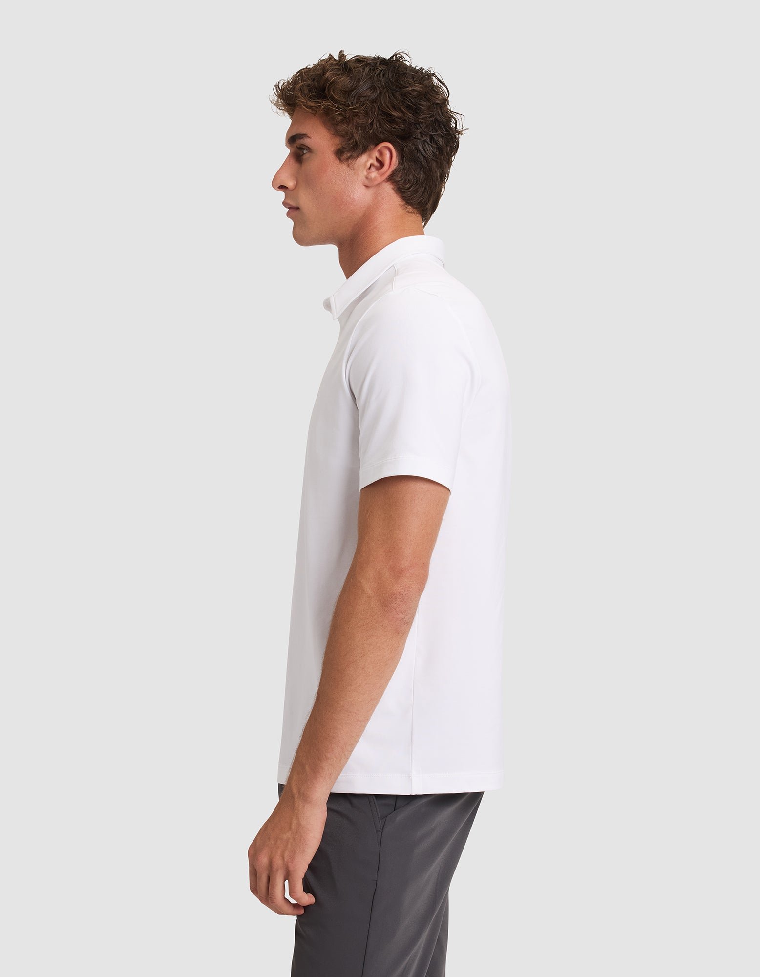 A young man stands in profile against a plain light gray background, wearing the Cozy Earth Men's Everyday Polo in white and gray pants. 