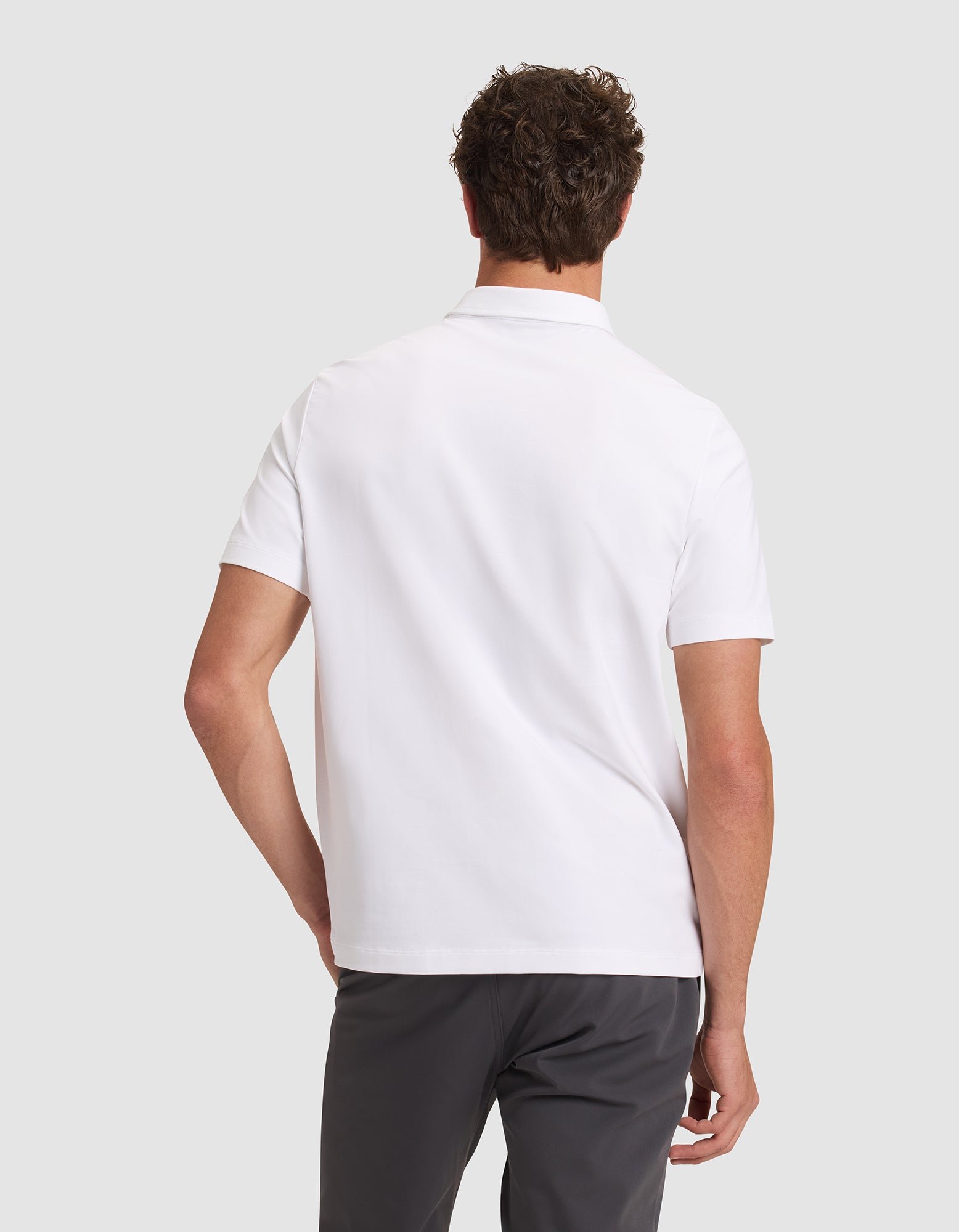 A man is seen from behind wearing the Cozy Earth Men's Everyday Polo in white with dark gray pants, standing against a light gray background. 