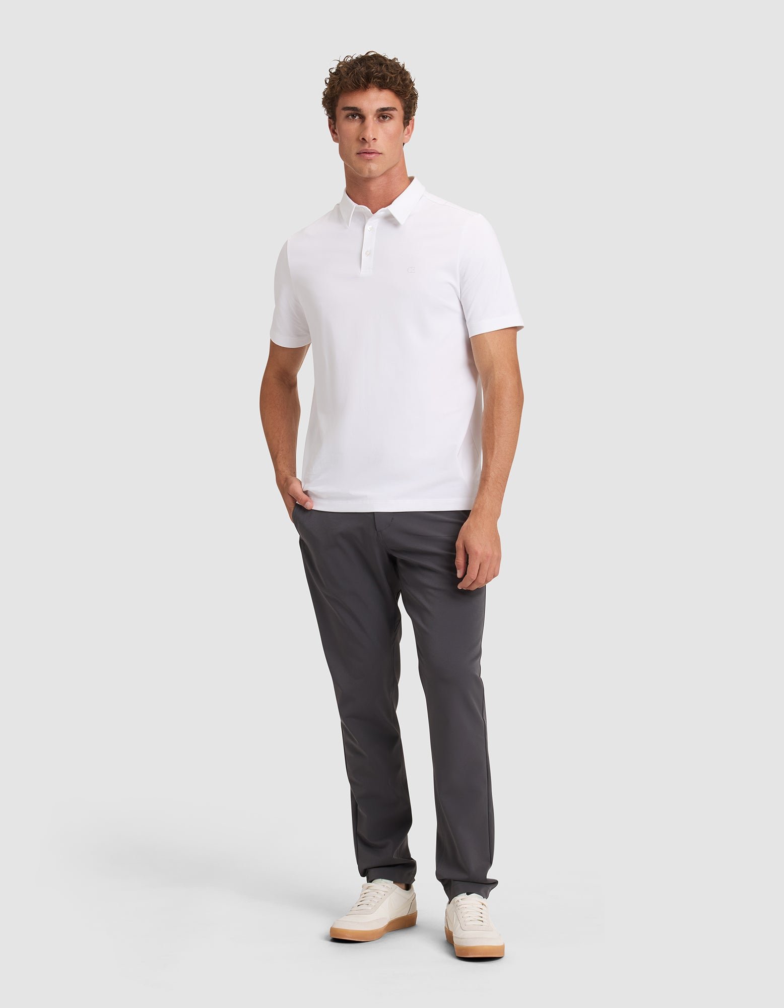 A man with short curly hair and one hand in his pocket models the Cozy Earth Men's Everyday Polo against a plain background, paired with dark gray pants and white sneakers. 