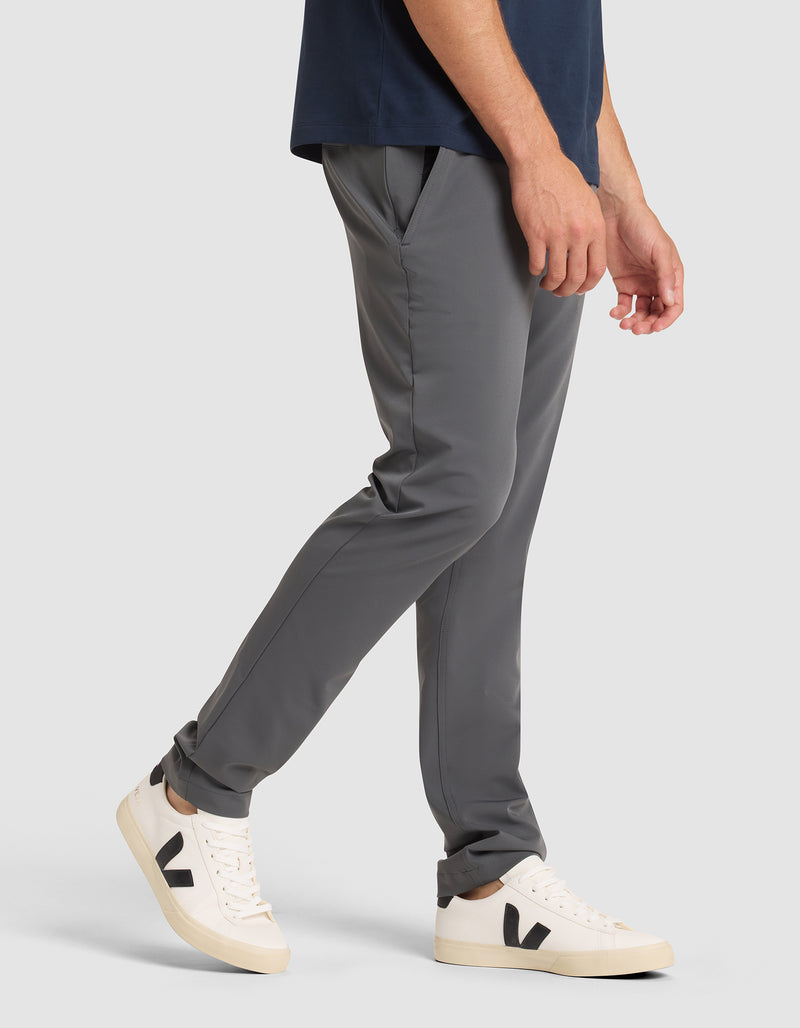 A man models the Cozy Earth Men's Everywhere Pant in gray with a navy shirt and white sneakers featuring black "V" logos, standing against a plain gray background, shown from shoulder to feet.